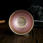 Handmade Jianzhan golden lotus tea cup with iridescent reddish-brown glaze and metallic gold lotus pattern - top-down close-up view on dark wooden surface