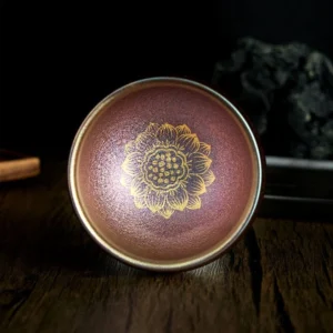 Handmade Jianzhan golden lotus tea cup with iridescent reddish-brown glaze and metallic gold lotus pattern - top-down close-up view on dark wooden surface