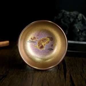 Handmade Jianzhan koi fish lotus ceramic tea cup with bronze-gold glaze and purple inner base, top-down flat view on wooden surface