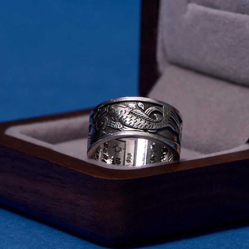 Koi fish heart sutra ring in antique silver resting on dark wooden jewelry gift box, 3/4 front angle showing carved fish and inner text
