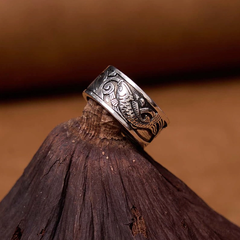 Koi fish sutra carved ring close-up on wooden display stand, aged silver patina showing intricate fish pattern carving