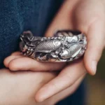 Koi fish lotus silver cuff bracelet with embossed prosperity symbols worn on wrist with blue linen
