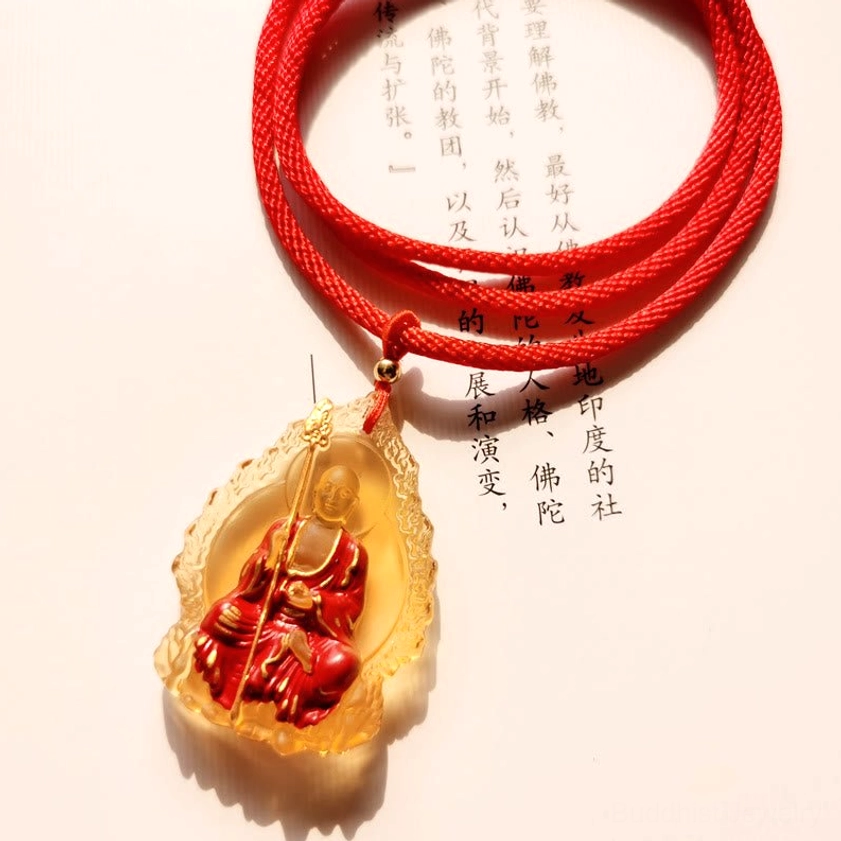 Amber translucent Ksitigarbha Buddha liuli crystal seated pendant on scripture paper background, oblique close-up with red braided necklace