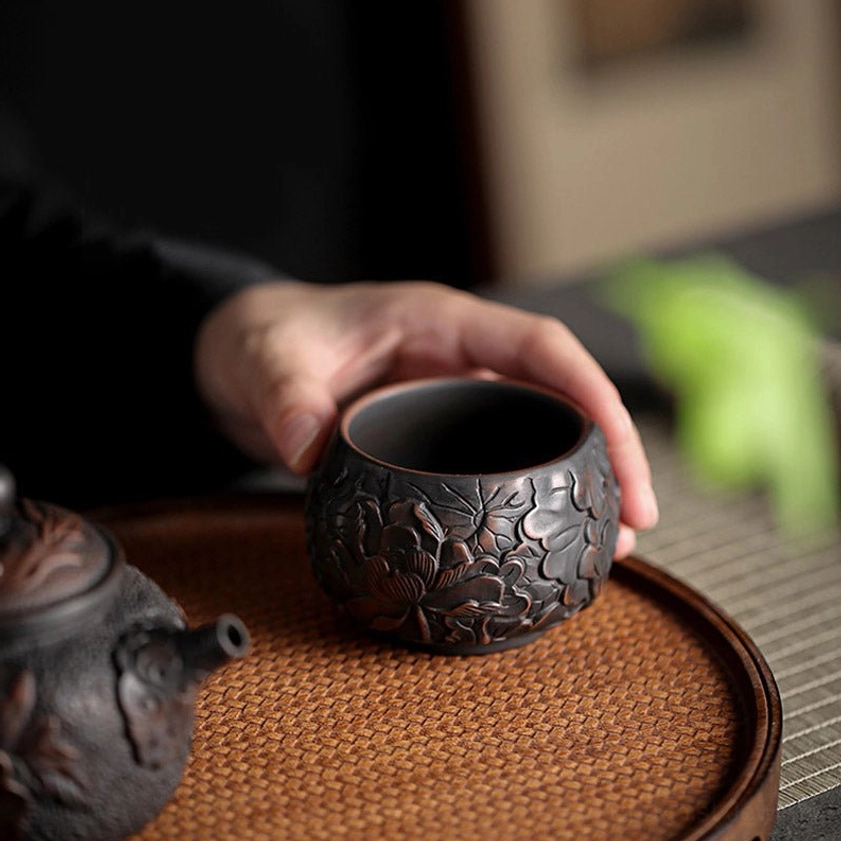 Handcrafted ceramic tea cup placed on woven tea tray next to matching teapot with embossed exterior details
