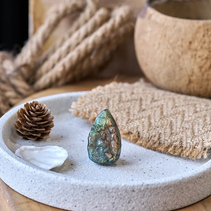 Mini labradorite Buddha carving displayed on decorative tray, front view showing blue-green iridescent sheen and carved Buddha relief
