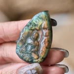 Labradorite Buddha teardrop statue with iridescent green-brown shimmer, hand-held close-up view showing carved seated Buddha relief and natural labradorescence