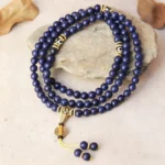 108 bead lapis lazuli Buddhist prayer mala with Dzi bead and gold accents displayed on natural stone with decorative leaves