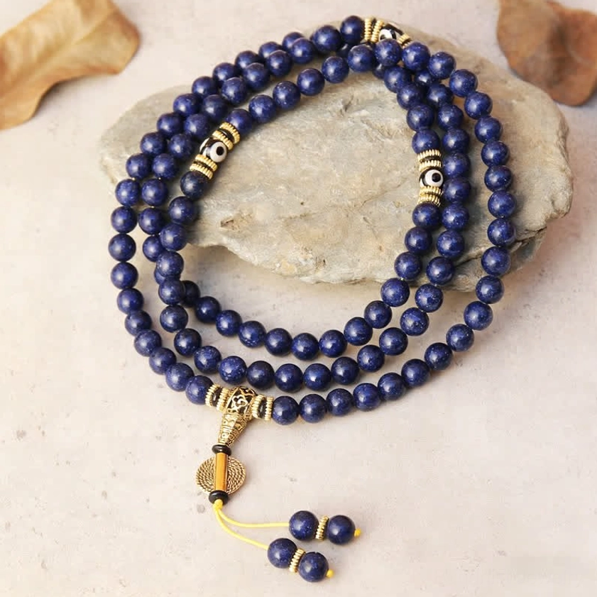 108 bead lapis lazuli Buddhist prayer mala with Dzi bead and gold accents displayed on natural stone with decorative leaves