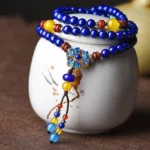Multi-layer Lapis Lazuli and Yellow Agate mala bracelet with blue enamel flower pendant, displayed on white ceramic jar for jewelry photography