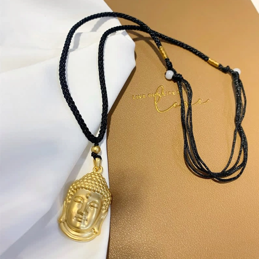Large gold Buddha head pendant necklace with black woven rope and white bead accents, top-down flat lay