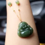 Laughing Buddha Hetian jade pendant necklace with 18K gold chain worn on wrist, showing detailed carved Buddha design and dark green jade stone
