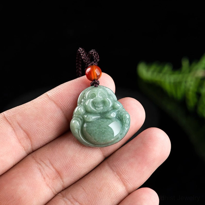 Miniature Laughing Buddha jade pendant held between fingers, showing intricate carving detail and polished green jade texture