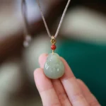 Laughing Buddha Hetian jade pendant held in palm, showing carved green jade with red accent bead and gold clasp detail