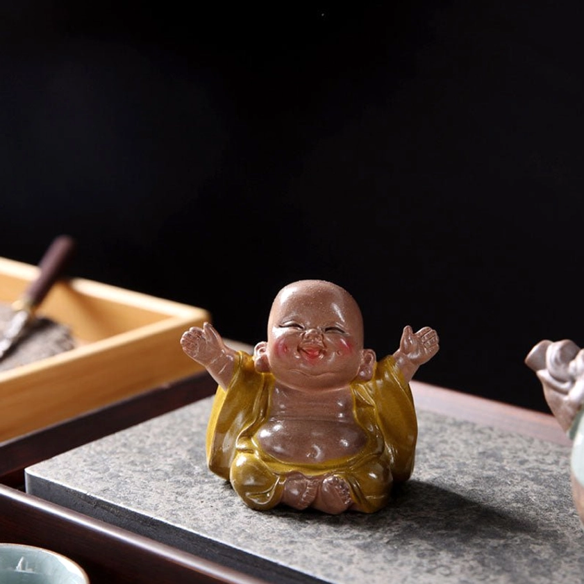 Top-down view of small laughing Buddha tea pet placed on stone tea mat with tea ceremony storage box in background