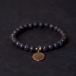 Lava rock beads bracelet with antique bronze copper pendant, three-quarter overhead view showing matte black porous volcanic beads and metal disc charm