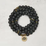 Lava rock 108 bead mala bracelet coiled flat lay with bronze lotus charm pendant on light gray background