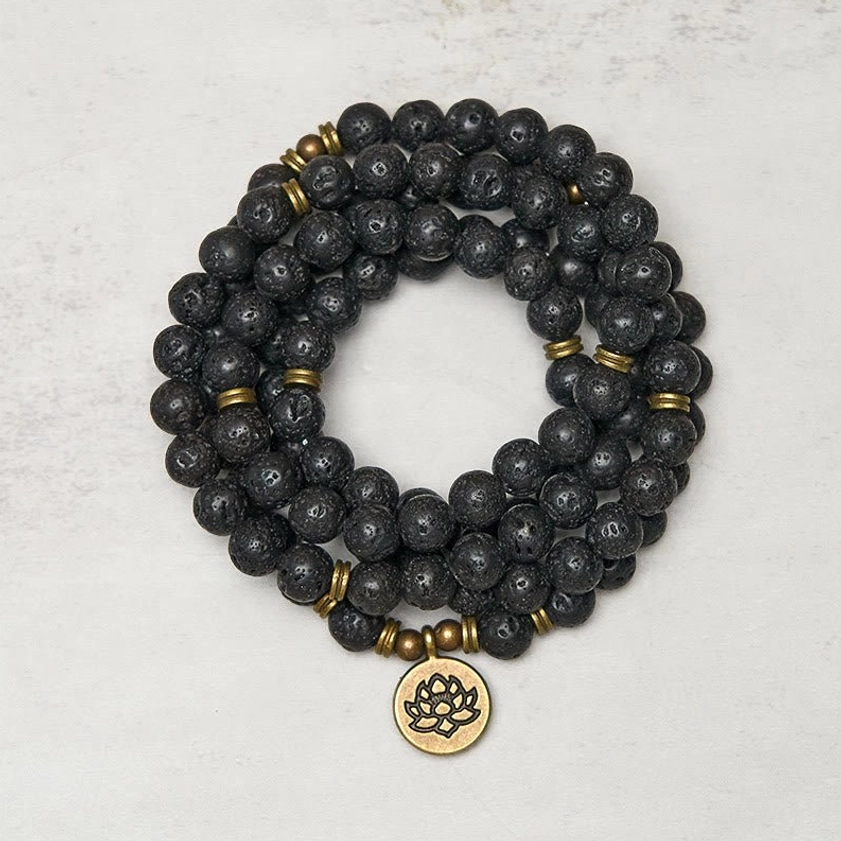 Lava rock 108 bead mala bracelet coiled flat lay with bronze lotus charm pendant on light gray background