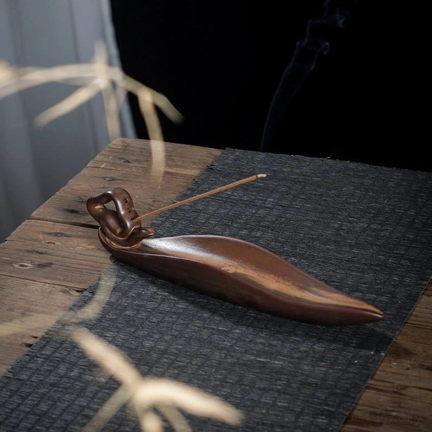 Dark matte ceramic leaf incense holder with twisted knot design and rising incense smoke on wooden desktop