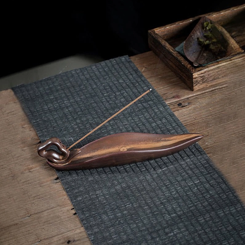 Leaf-shaped ceramic incense burner with twisted knot end holding incense stick on wooden table with woven mat