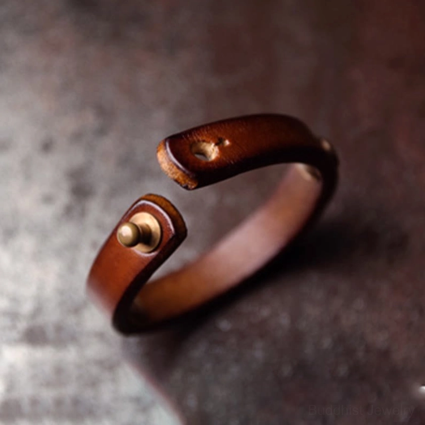Polished leather and brass bangle strap end with exposed brass rivet on textured dark surface