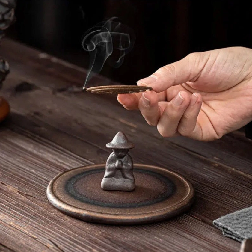 Hand lighting incense stick in kneeling monk ceramic incense holder on wooden tea table for meditation and mindfulness