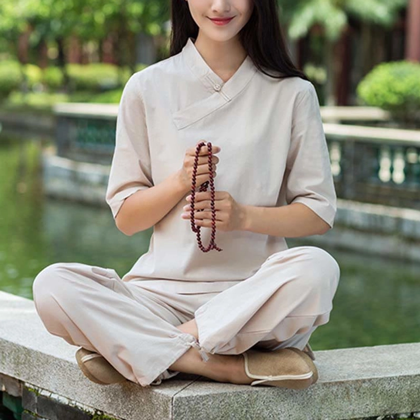 Woman wearing light khaki linen meditation outfit with cross V-neck design and half sleeves, standing by water in zen garden