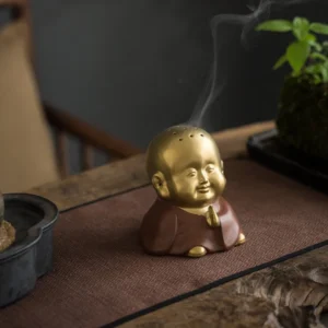 Little Buddha ceramic incense burner front view with praying hands on wooden tea table, matte gold and brown finish with rising incense smoke