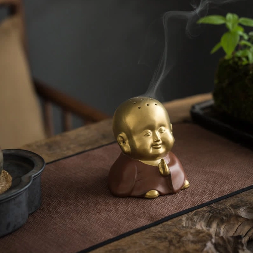Little Buddha ceramic incense burner front view with praying hands on wooden tea table, matte gold and brown finish with rising incense smoke