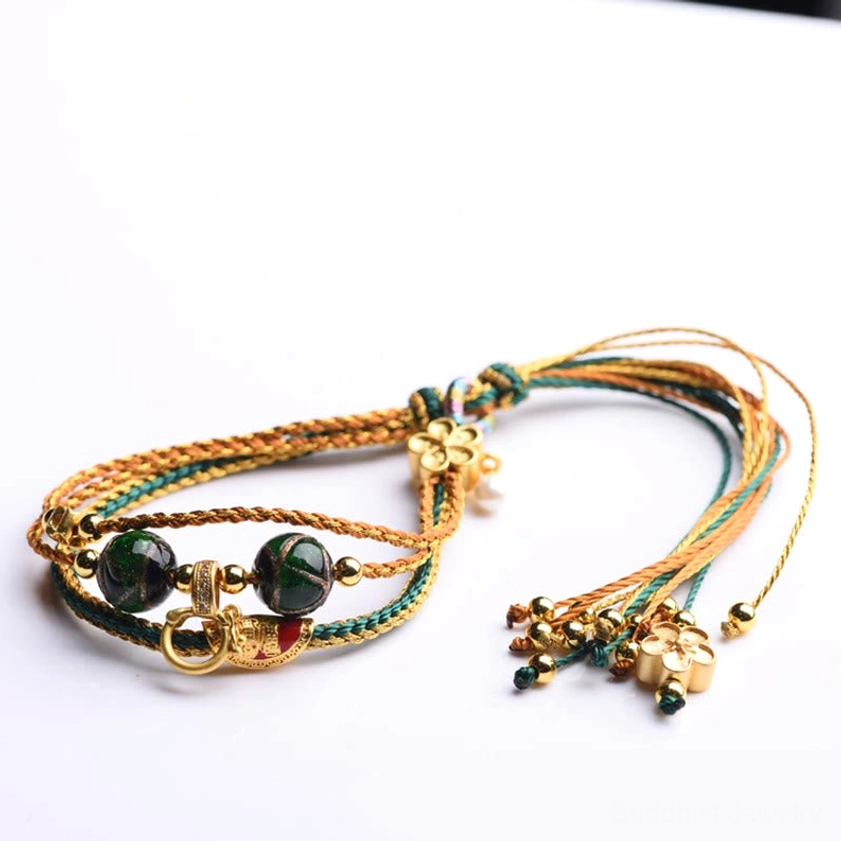 Flat-laid handmade liuli glass bracelet featuring gold-teal woven cord, dark green glass beads, tassel pendants, and metal ring accents