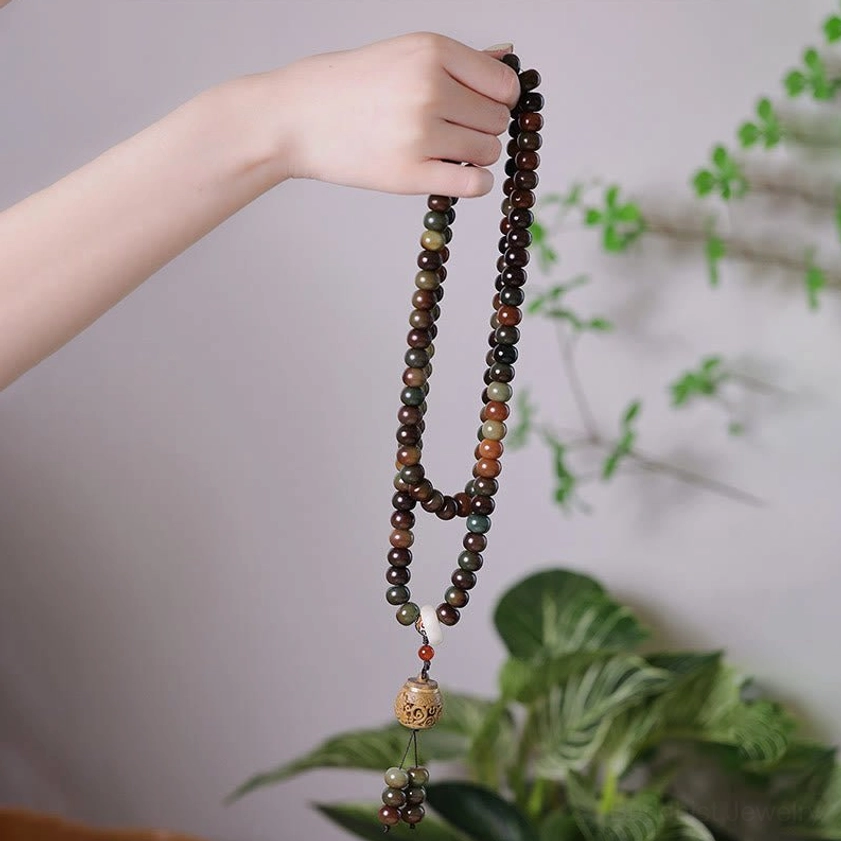 Long mala prayer beads necklace held in hand with green plant decor background, showing earthy brown beads and wooden pendant