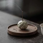 Handcrafted white ceramic lotus incense burner with burning incense stick on dark wooden tea table with round wooden tray
