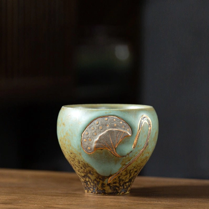 Turquoise lotus pod engraved ceramic tea cup with gold embossed design, frontal studio view on wooden table