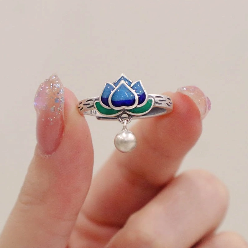 Lotus pattern enlightenment ring held by manicured hand displaying open adjustable band, blue cloisonné lotus, and silver bell pendant
