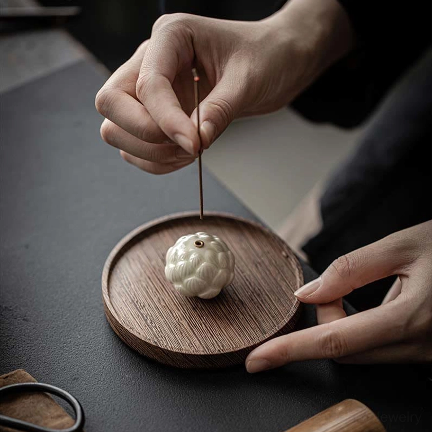 Hands placing incense stick into ceramic lotus holder on dark desktop with wooden tray and scissors nearby