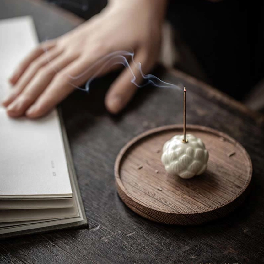 Ceramic lotus incense holder with wispy aromatic smoke rising, hand resting nearby on desktop with notebook