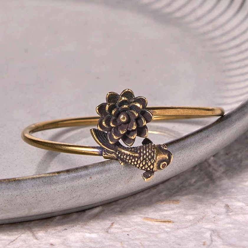 Vintage antiqued brass lotus koi bangle close-up detail shot on light grey ceramic background showing carved lotus and koi fish charm intricately