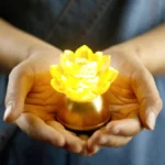 Warm glowing yellow lotus liuli glass prayer light with rose-gold copper base held in hands, close-up handheld view