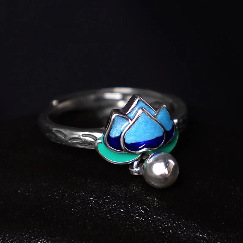 Adjustable lotus enamel ring showing engraved silver band with blue-green lotus design and silver bell charm in flat lay composition