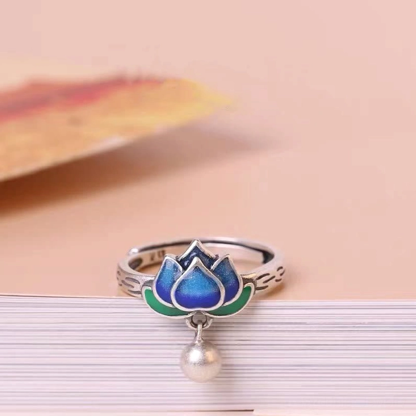 Exquisite lotus ring placed on stacked books showing smooth silver band, white pearl pendant, and vibrant blue-green enamel lotus