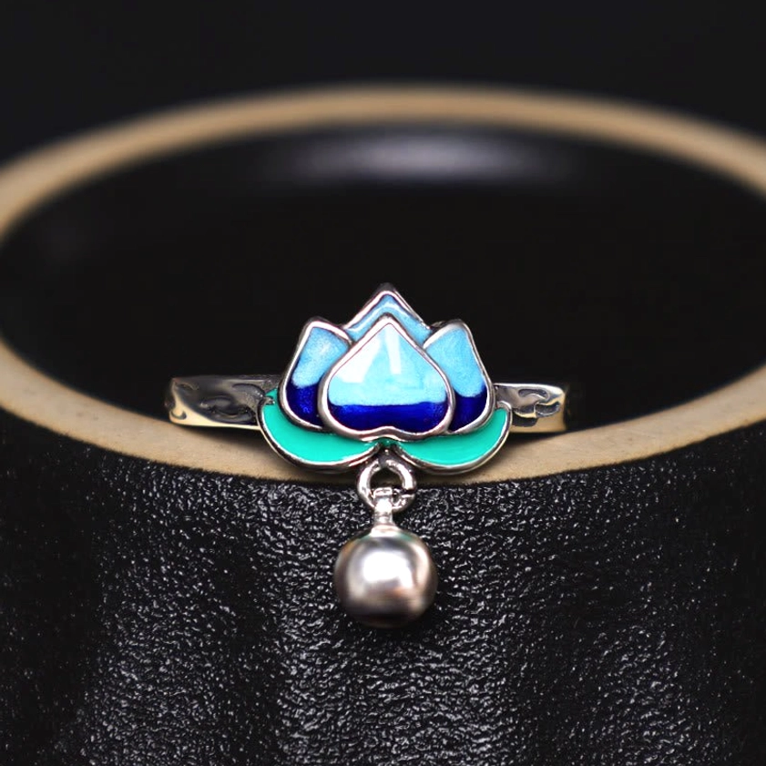 Buddhist lotus silver ring with blue-green enamel and dangling bell accent displayed on black leather vessel rim, side angle