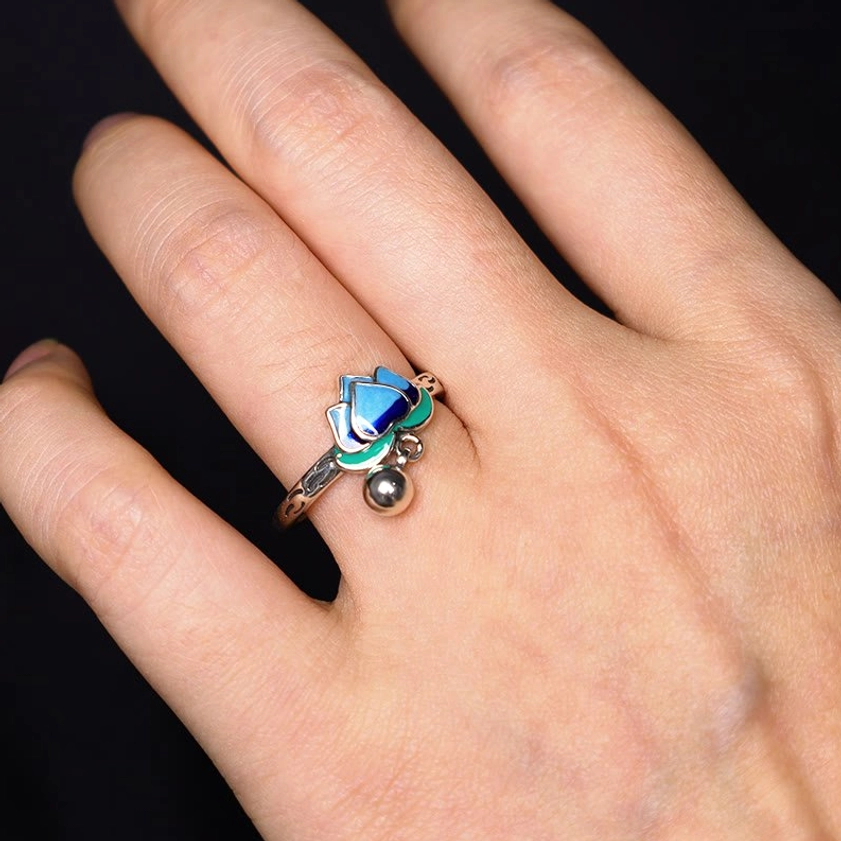 Buddhist lotus ring worn on woman's finger showing engraved silver band, blue-green enamel design, and dangling bell charm