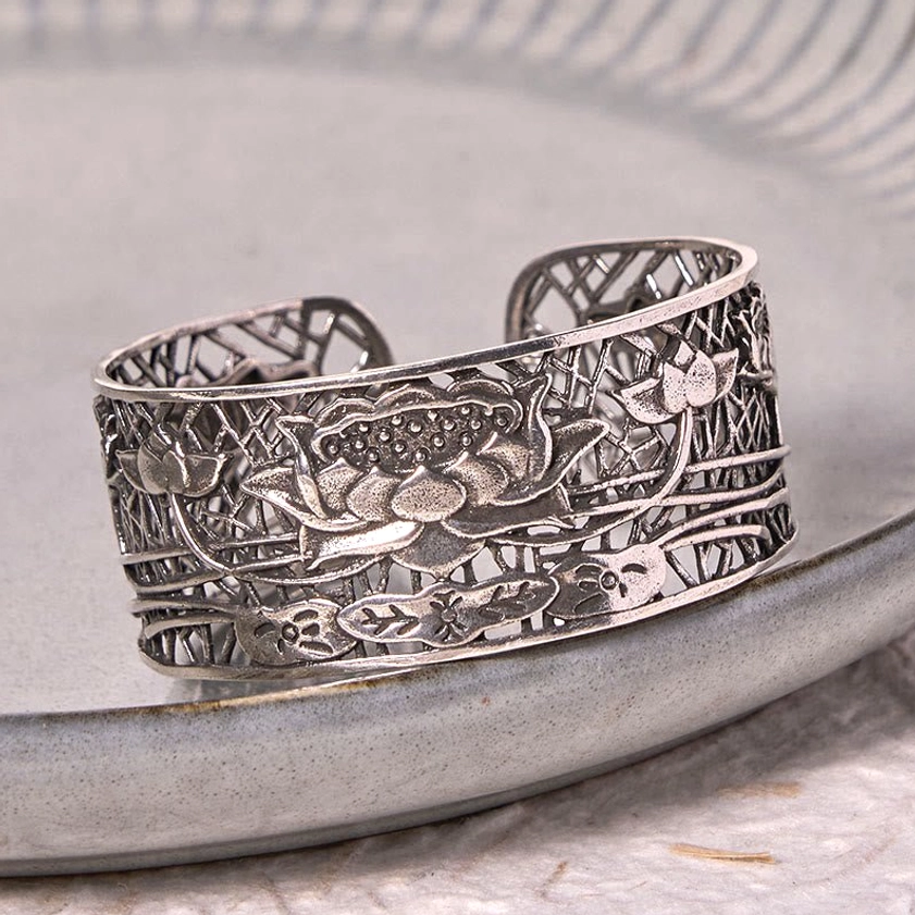 Silver lotus relief cuff bracelet three-quarter view on light grey ceramic tray, displaying antique metallic tone and carved floral details