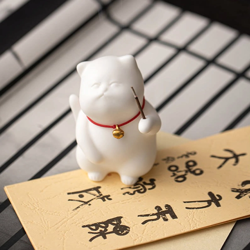 White ceramic lucky cat figurine on vintage greeting card backdrop with incense stick, side view