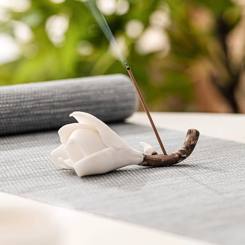 Lit incense burning in ceramic magnolia holder with rising fragrant smoke, placed on grey linen mat with natural green foliage background