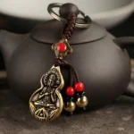 Mahasthamaprapta Buddha gourd blessing keychain with bronze finish displayed on dark clay teapot, showing embossed Buddha design and red bead decorations with golden bell pendants