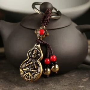 Mahasthamaprapta Buddha gourd blessing keychain with bronze finish displayed on dark clay teapot, showing embossed Buddha design and red bead decorations with golden bell pendants