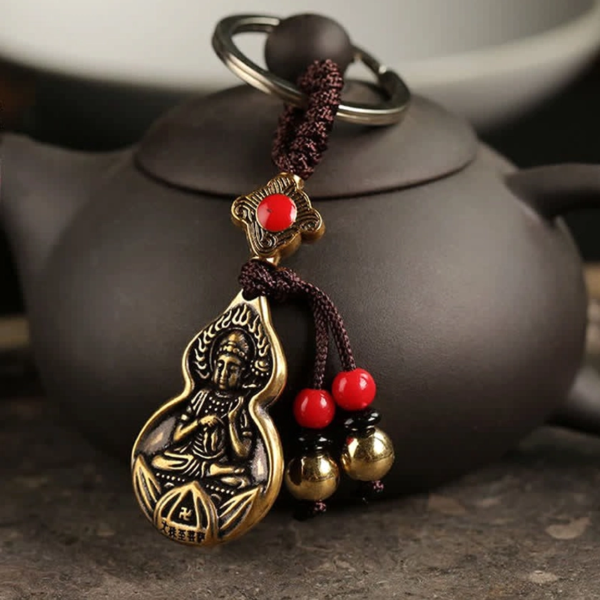 Mahasthamaprapta Buddha gourd blessing keychain with bronze finish displayed on dark clay teapot, showing embossed Buddha design and red bead decorations with golden bell pendants