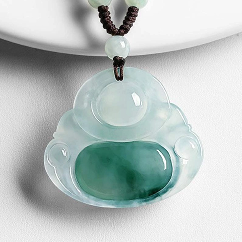 Light cyan jade Maitreya Buddha pendant with smooth carved Buddha figure, green streaks in jade stone, and brown decorative cord on white ceramic surface