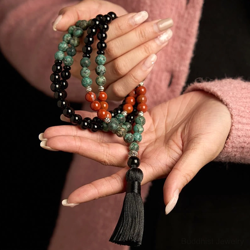 Hands holding 108-bead mala bracelet showing detailed view of mixed gemstone beads, black tassel charm, and artisan craftsmanship
