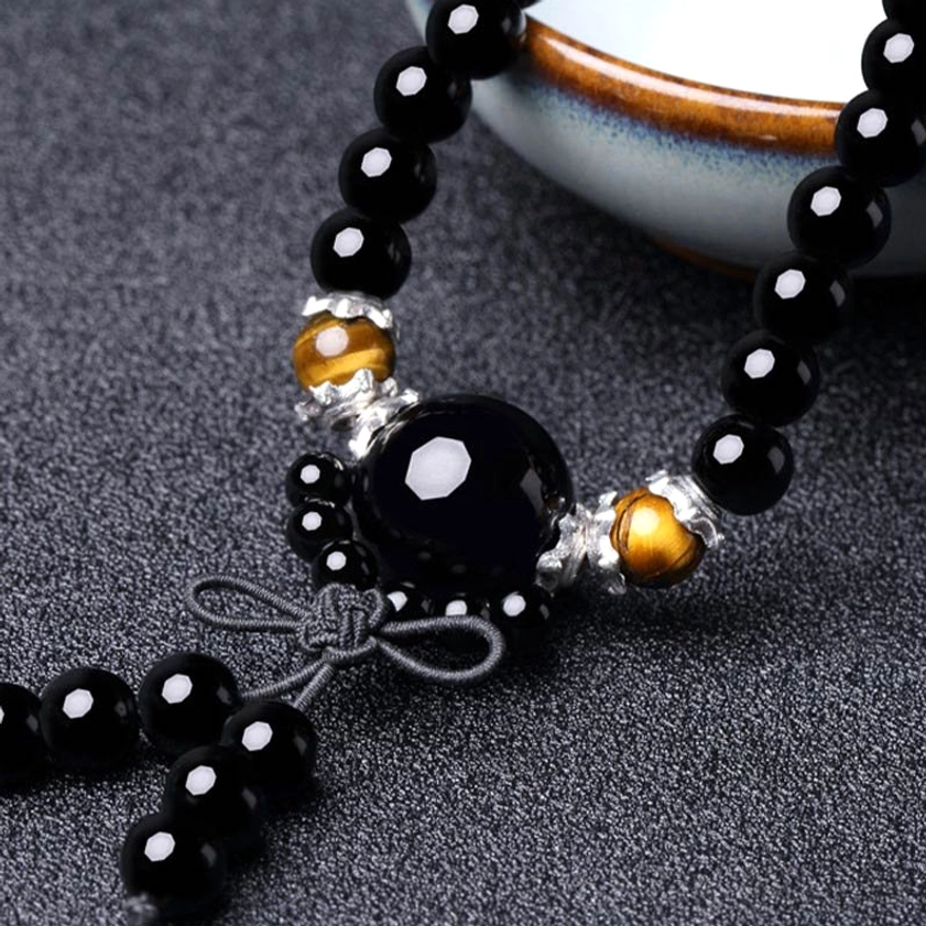 Macro close-up of black obsidian tiger eye Buddhist mala bracelet with silver wrap and knotted tassel beside ceramic tableware